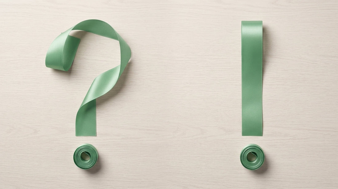Two identical sage green satin ribbons arranged on a minimalist wooden table, one curled into a question mark and the other into an exclamation mark, representing a quality comparison study for Peaco ribbons.